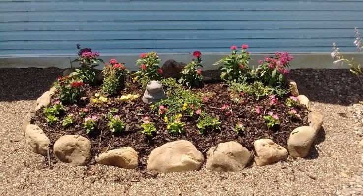 A circular flower bed bordered with large natural rocks, set on gravel against a light blue slatted wall